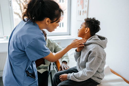 Female pediatrician checking neck ache of boy sitting on examination table with father in medical room