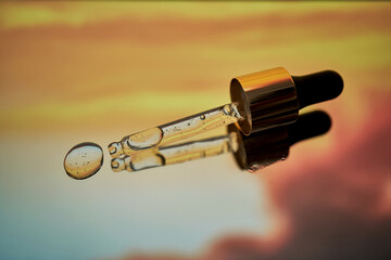 A pipette with facial serum against a sunset background in bright orange tones.