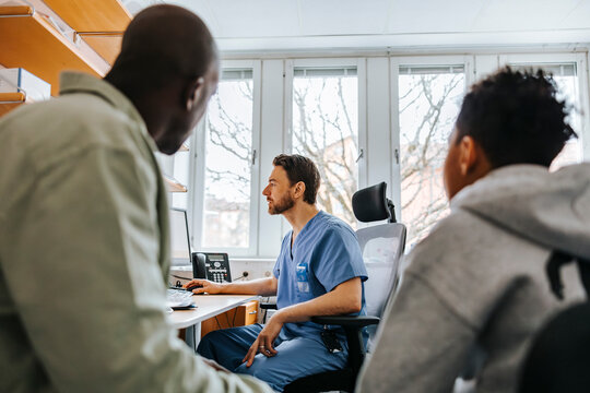 Male medical expert in scrubs working on computer while sitting with patient's in examination room