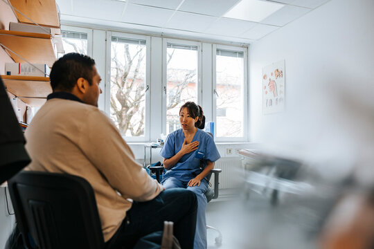 Female doctor with hand on chest talking with male patient while sitting in medical examination room