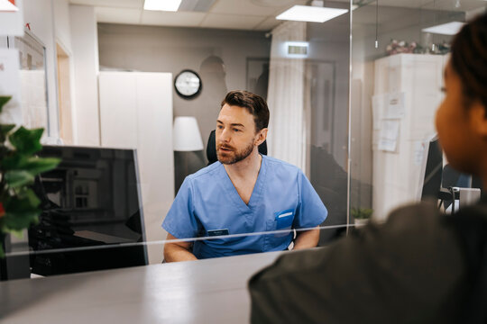 Male receptionist looking at computer and assisting patient standing at hospital