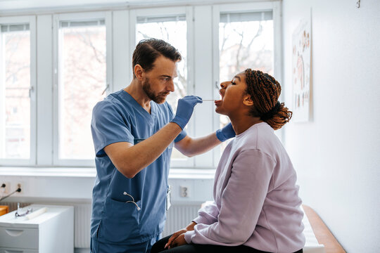Male medical expert examining throat of woman using tongue depressor in examination room at hospital
