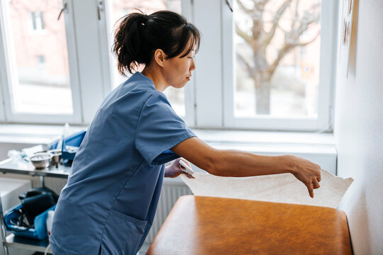 Side view of female healthcare worker putting sheet on bed in examination room