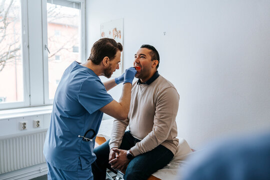 Male medical expert examining throat of man using tongue depressor in examination room