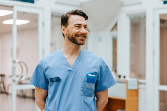 Happy male doctor wearing scrubs looking away with hands behind back in hospital