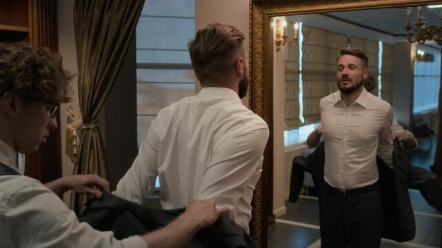 Man trying on new elegant suit with help of salesman and looking at his reflection in mirror while visiting clothing store