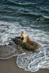 A beautiful blonde girl sits on a stone among the waves on the beach, on a sunny summer day.