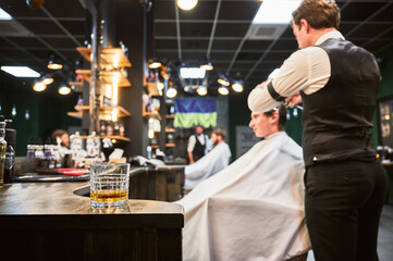 Close-up photo of a glass of whiskey. High-quality barbershop offering drinks to customers. Glass with alcohol on the background of hairstylist making haircut.