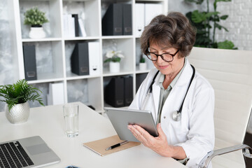 Elderly senior female doctor pediatrician general practitioner using digital tablet for consulting patients online, remote telemedicine at hospital