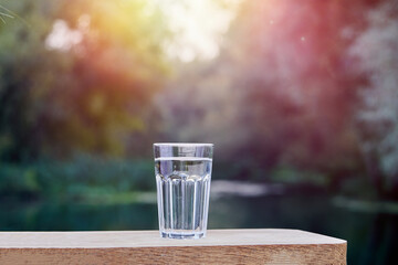 Glass of fresh water is standing on a wooden surface outdoors, with a peaceful river flowing in the background