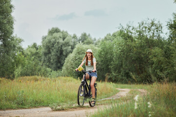 Fototapeta premium Young happy woman in a casual wear rides a bicycle in summer meadow. Beautiful girl enjoys summer nature. Relax, rest, nature concept.