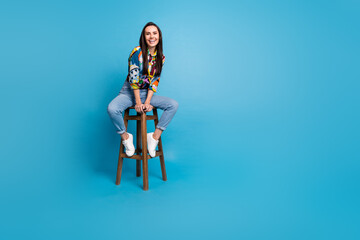 Naklejka premium Full length photo of pretty nice pleasant woman wear print shirt denim trousers sitting on bar stool isolated on blue color background