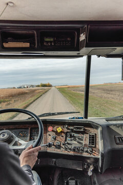 Inside View of an Old Semi Truck Dashboard: Dusty and Worn with Use, Featuring the Driver&rsquo;s Hand on the Steering Wheel as He Shifts Gears, Driving on a Rural Dirt Road with Corn Crops and a Blue Sky