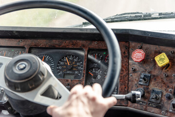 Inside View of an Old Semi Truck Dashboard: Dusty and Worn with Use, Featuring the Driver’s Hand on the Steering Wheel as He Shifts Gears, Driving on a Rural Dirt Road with Corn Crops and a Blue Sky
