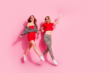 Full length photo of two lovely young ladies jump running hold hands dressed stylish red leopard print garment isolated on pink background