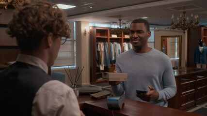 African American customer paying by mobile phone, taking package from cashier and thanking him while making purchase at counter in menswear store