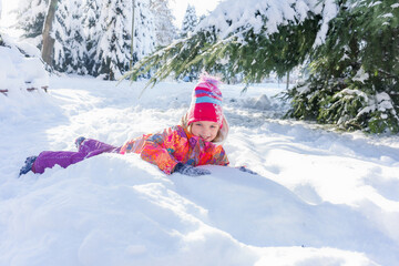 Portrait of a happy 6-year-old girl in a colorful winter suit lying in deep snow in the park. Children's winter fun.