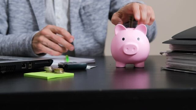 In an office setting, a man diligently places a banknote into a pink piggy bank, managing his finances and organizing his savings on the desk