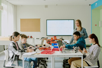 Obraz premium Multiracial students sitting at work desk and creating robots while their teacher standing next to table and watching them
