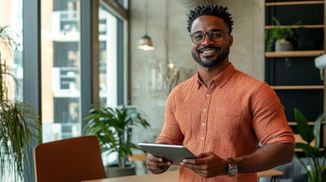 A black man with short, curly hair is wearing an orange button-down shirt and glasses. He smiles at the camera as he holds a tablet in front of him. There are plants and windows in the background.