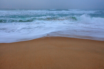 Gentle waves roll towards the sandy shore under a misty, overcas