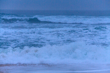 Layers of powerful waves roll towards the shore in a misty, blue-tinted scene, capturing the energy of the ocean