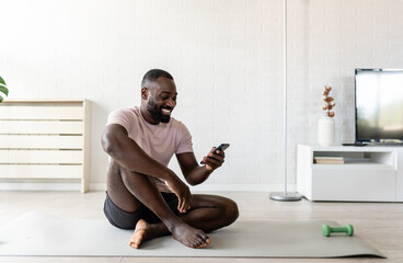 A young man sits on a yoga mat at home after exercising, smiling and using his smartphone, surrounded by plants and natural light in a relaxed atmosphere.