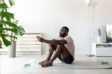 A young African-American man sits on a yoga mat at home after exercising, smiling and using his smartphone, surrounded by plants and natural light in a relaxed atmosphere.