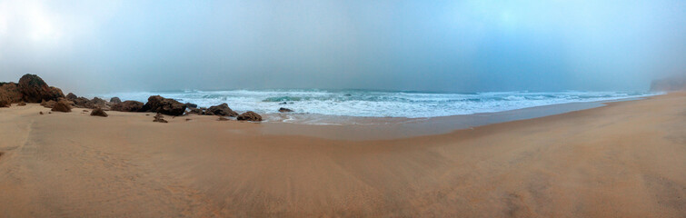 A foggy beach scene featuring scattered rocks along the sandy sh