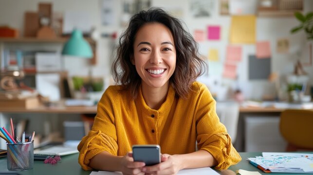 A cheerful woman in a mustard sweater holds a smartphone while sitting in a cozy, creatively cluttered workspace filled with colorful notes and art supplies.