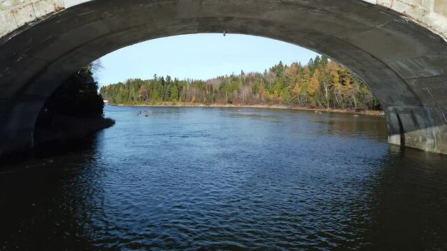 Scenic drone footage gliding above the Mitis salmon river and passing beneath the Arthur‑Bergeron Bridge arch as it flows toward its mouth under clear blue skies on Quebec&rsquo;s Gasp&eacute; Peninsula.