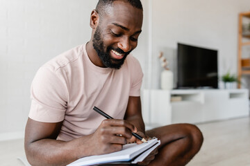 Young African-American man sitting cross-legged on a yoga mat at home, writing in a notebook after...