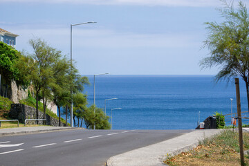 Empty coastal road descending towards a calm blue ocean on a clear day, bordered by trees and...