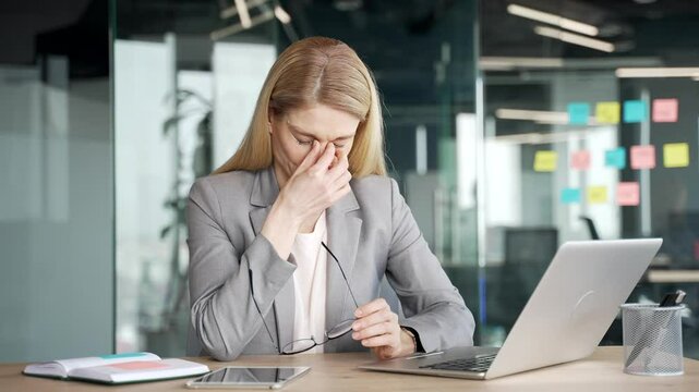 Tired businesswoman in suit suffering from eye strain and headache while working on laptop sitting at desk in business office. Overworked woman takes off glasses and massages, rubs bridge of nose