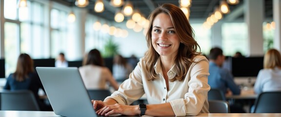 A confident young woman smiling while working on a laptop in a modern office environment. The setting is bright and collaborative, fostering a sense of productivity and teamwork.

