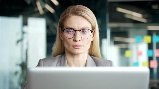 Serious thoughtful businesswoman works on laptop sitting at workplace in business office. Female employee in glasses is busy with a project, banking, texting or chatting online. Front view. Close up