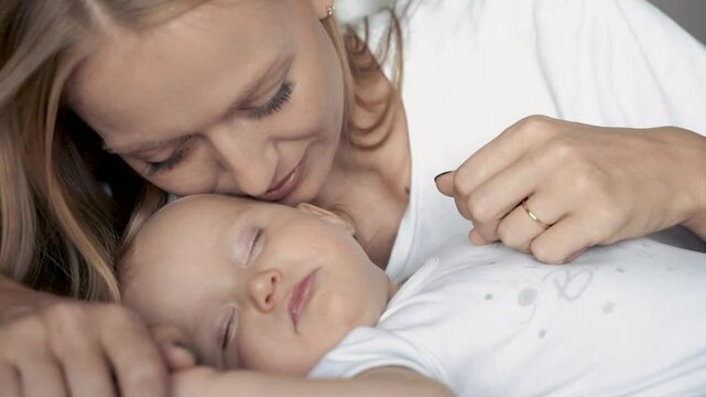 Happy mom touching and kissing sleeping newborn baby. Cute newborn baby sleeping in bed at home. Family care, happy childhood and motherhood, close up