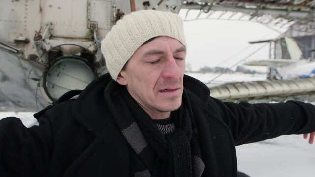 A Caucasian man with gray eyes who is desolated in sadness and raises his hands and looks up in despair and throws his head back. In the background is an old airport with broken planes. Winter scene.