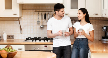 Happiness At Home. Cheerful young family of two drinking hot tea standing in modern kitchen, looking at each other