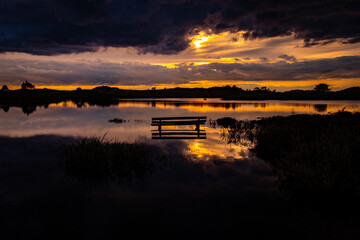 Solitary Bench Reflecting Sunset Glow Over a Tranquil Lake: Dramatic Sky and Still Waters Capture Nature’s Serene Beauty in Twilight’s Embrace