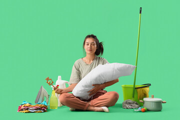 Overwhelmed Asian mother meditating with baby, toys, cleaning supplies and clothes on green...