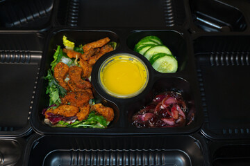 A salad in a black plastic container surrounded by catering containers, featuring fresh greens, vegetables, and sauce. The background of recyclable containers.