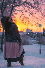 A woman wearing a stylish pink skirt is standing gracefully in the snow during a beautiful sunset, creating a stunning scene