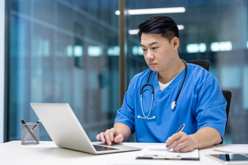 Serious asian doctor in blue scrubs using laptop and taking notes at desk within contemporary...