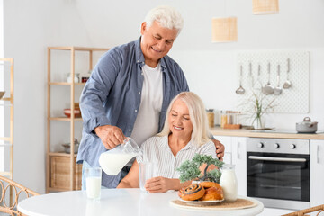 Happy senior couple with tasty buns pouring milk from jug into glasses in kitchen