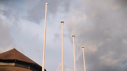 Four tall flagpoles stand empty under a cloudy sky, positioned beside a building with an octagonal...