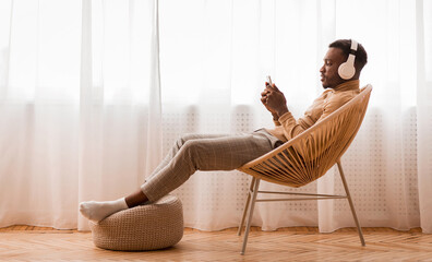 Weekend. Relaxed Black Man In Wireless Headset Using Mobile Phone Listening To Audiobook Sitting On...