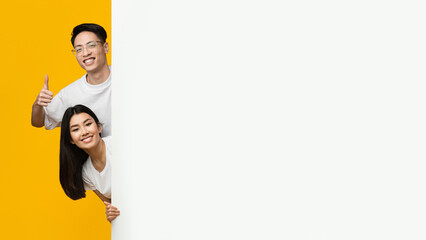 Happy asian young couple standing next to empty advertising board over yellow background, guy...