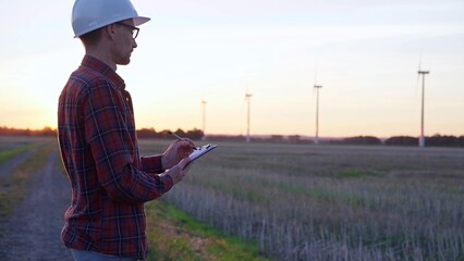Man engineer wearing a white protective helmet is taking notes with a clipboard in a field with wind turbines, as the sun sets. Clean energy and engineering concept