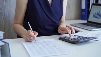 Businesswoman with dark blue dress is using dark calculator and taking notes of company expenses in her office. Taxes and audit in business
